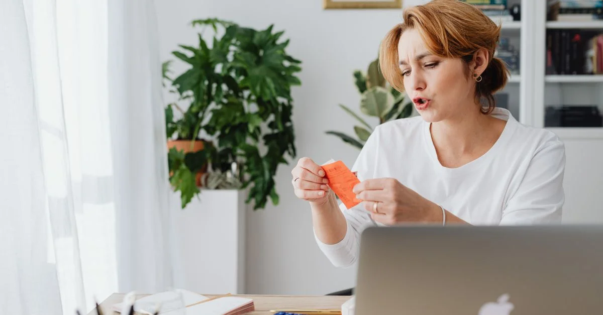 Mujer revisando facturas en el escritorio de casa con laptop y plantas, gestionando finanzas personales.
