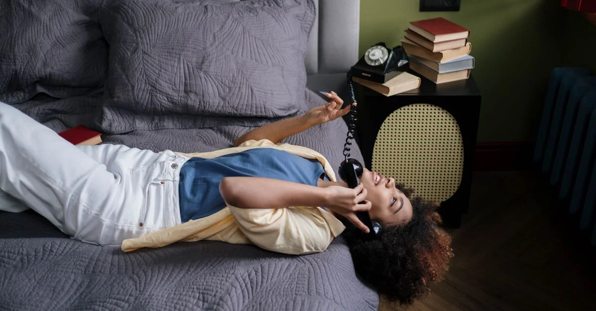 Joven mujer tumbada en la cama haciendo una llamada telefónica con un teléfono de disco vintage en casa.