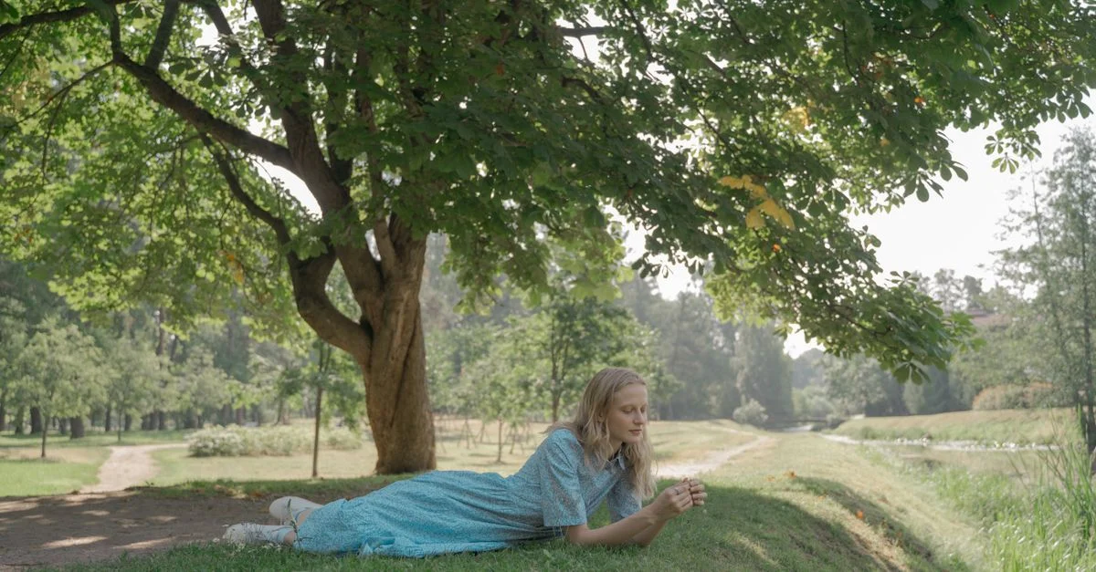 Una joven en un vestido azul acostada bajo un árbol en un día soleado en el parque, disfrutando del aire libre.