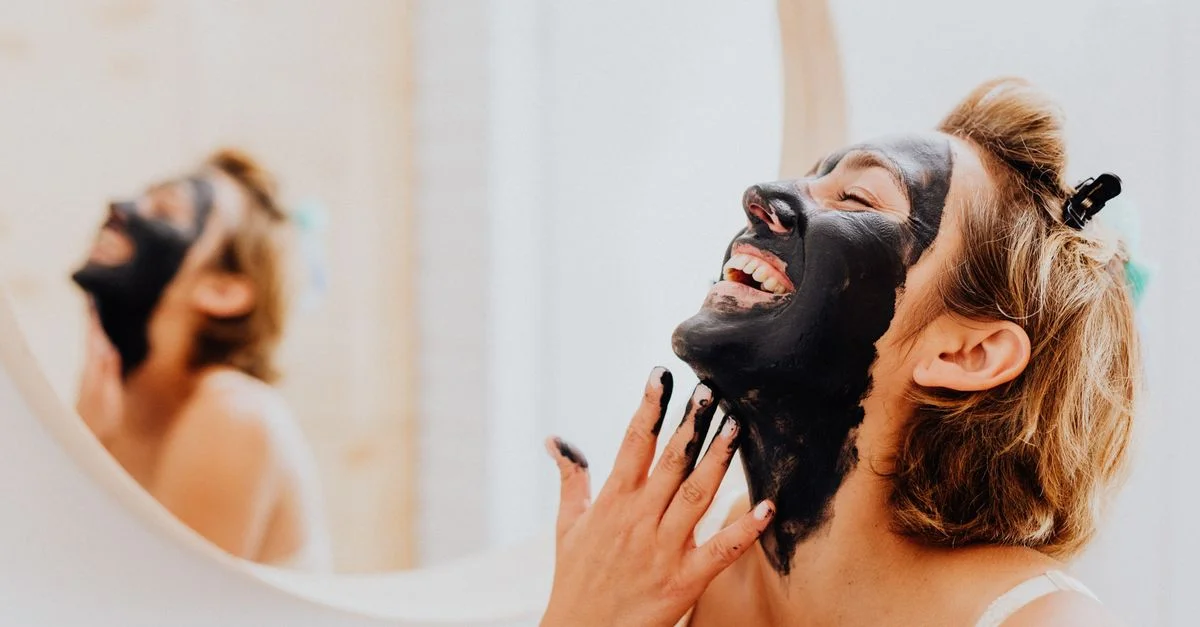 Una mujer alegre aplicándose una máscara facial negra mientras ríe en el baño.