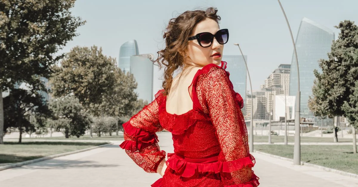 Una mujer elegante con un vestido rojo y gafas de sol posa con confianza al aire libre, con edificios modernos de fondo.