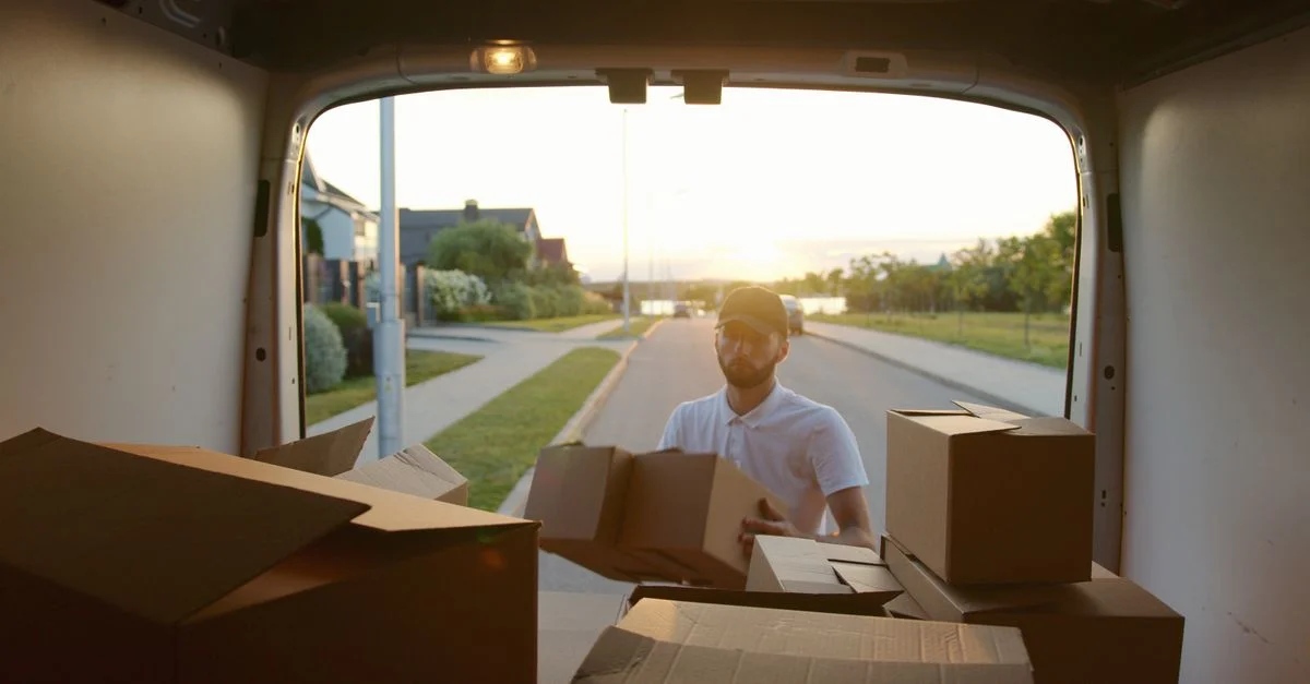 Un mensajero descargando cajas de cartón de una furgoneta durante el atardecer en una calle suburbana.
