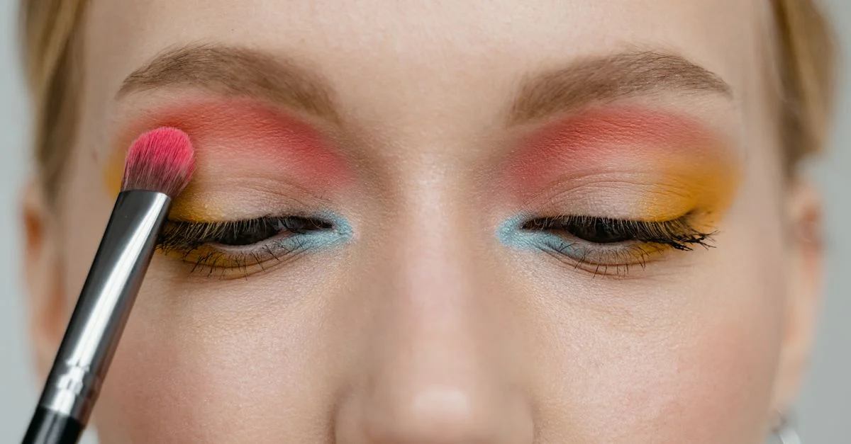 Primer plano de maquillaje de ojos vibrante con un pincel aplicando colores rosa, azul y amarillo.