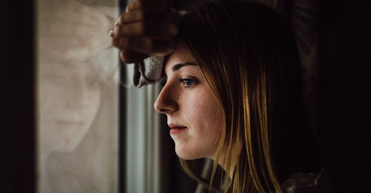 Una joven contemplativa mira por la ventana con una expresión reflexiva, en el interior.