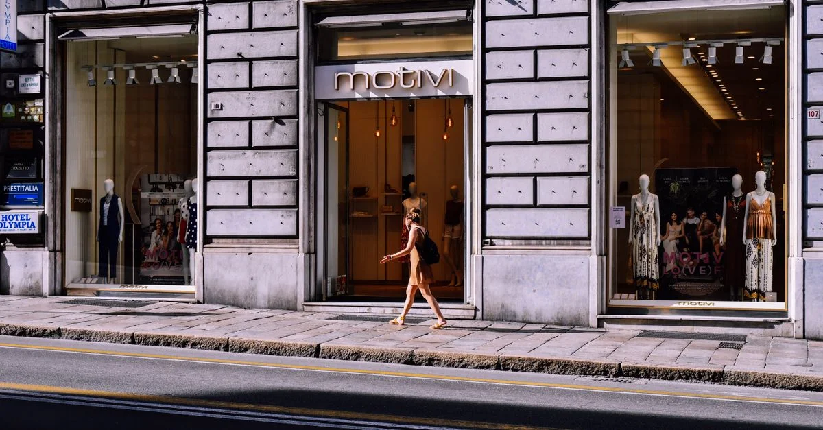 Mujer pasa junto a una tienda de ropa elegante en la acera urbana a la luz del día.