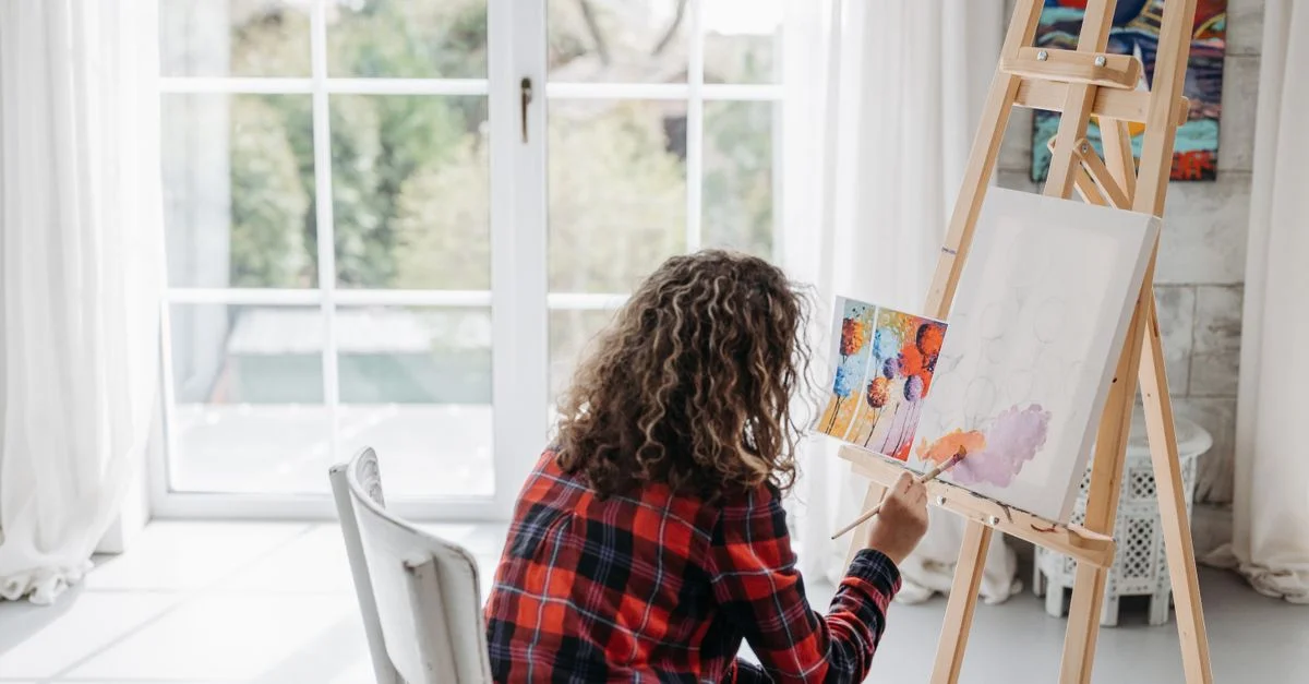 Una persona pintando en un lienzo en un estudio brillante y soleado con grandes ventanas.