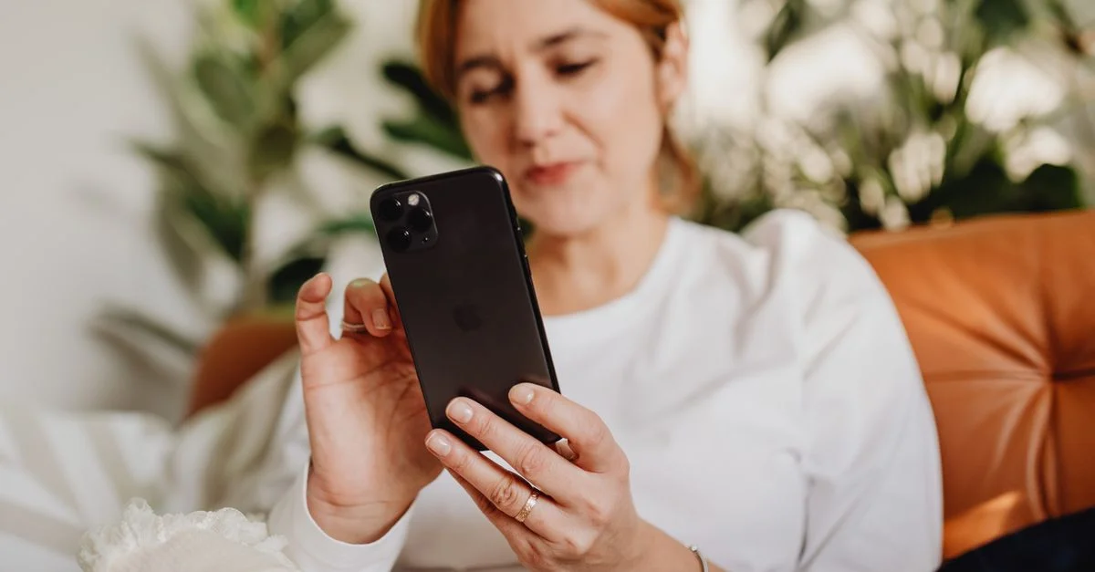 Mujer sentada cómodamente en interiores usando un teléfono inteligente con plantas de fondo.