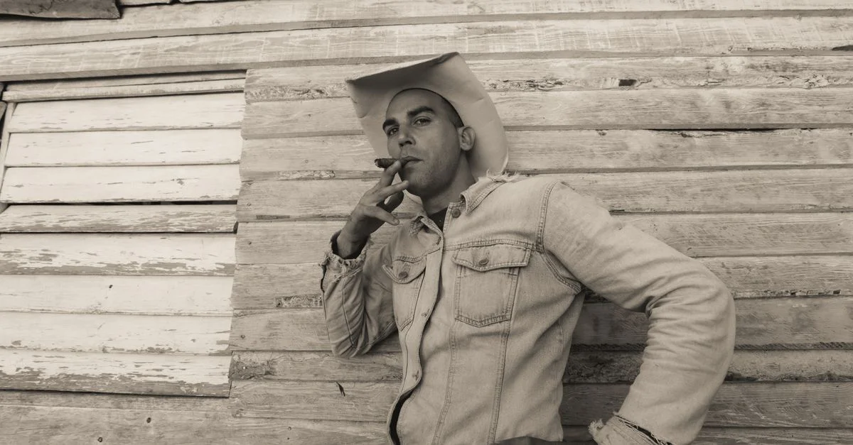 Un hombre con sombrero vaquero y chaqueta de mezclilla fuma un cigarro, apoyado contra una pared de madera.