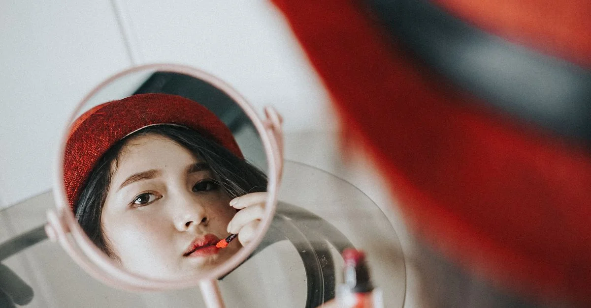 Primer plano de una mujer maquillándose los labios frente a un espejo, usando un sombrero rojo.