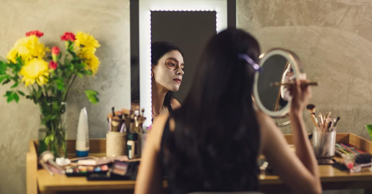 Una mujer aplicando pintura facial para el Día de los Muertos, reflejada en un espejo con un vibrante arreglo floral cerca.