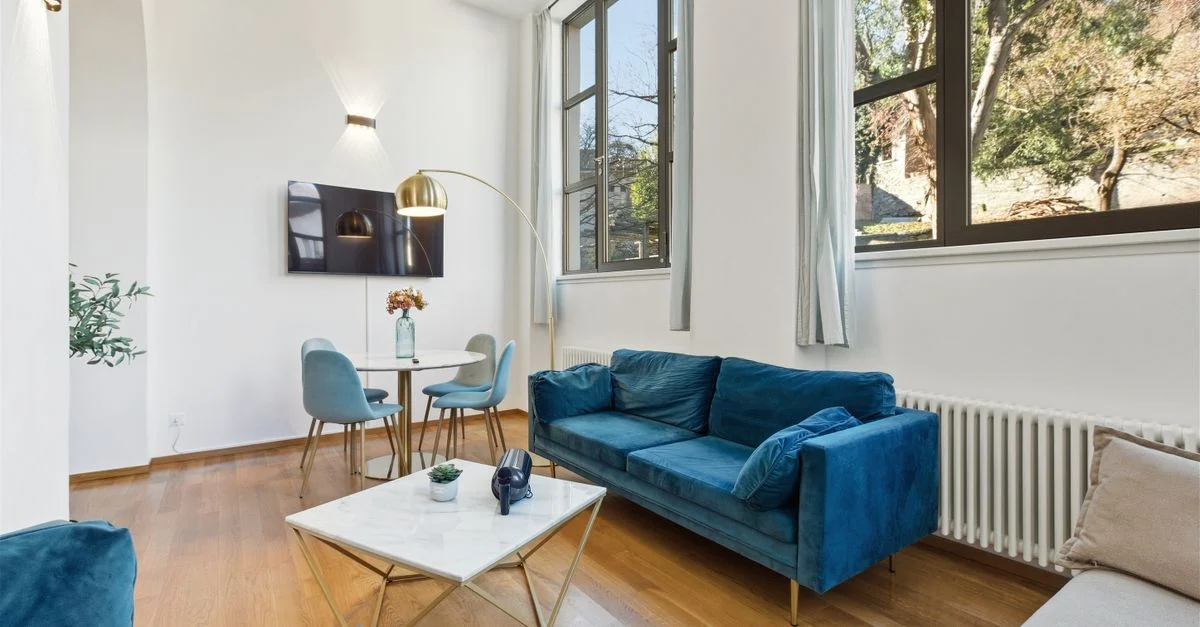 Salón luminoso y elegante con sofá de terciopelo azul, grandes ventanales y decoración minimalista.