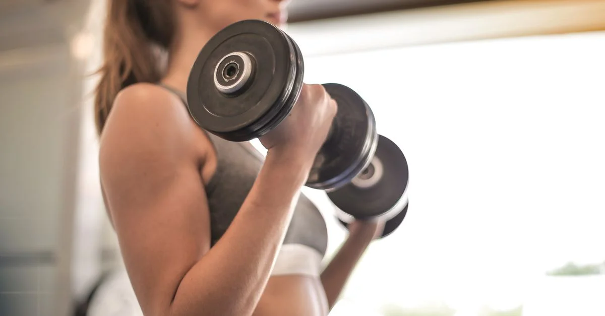 Mujer concentrada levantando mancuernas en un gimnasio brillante, enfatizando fuerza y fitness.