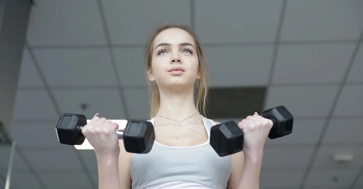 Joven mujer concentrada levantando mancuernas en un gimnasio interior.