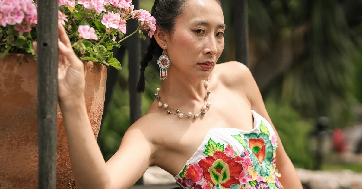 Una mujer elegante con un colorido vestido bordado posa al aire libre junto a flores rosas en plena floración.