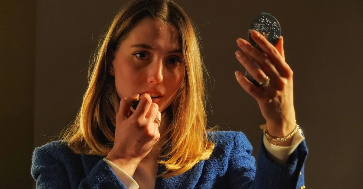 Mujer aplicándose lápiz labial mientras sostiene un espejo compacto en un entorno cálido y iluminado.