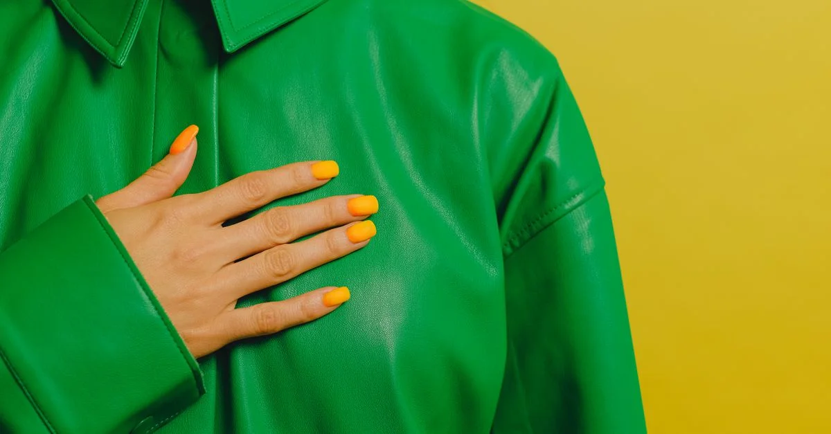 Primer plano de la mano de una mujer con uñas amarillas sobre una chaqueta de cuero verde contra un fondo amarillo.