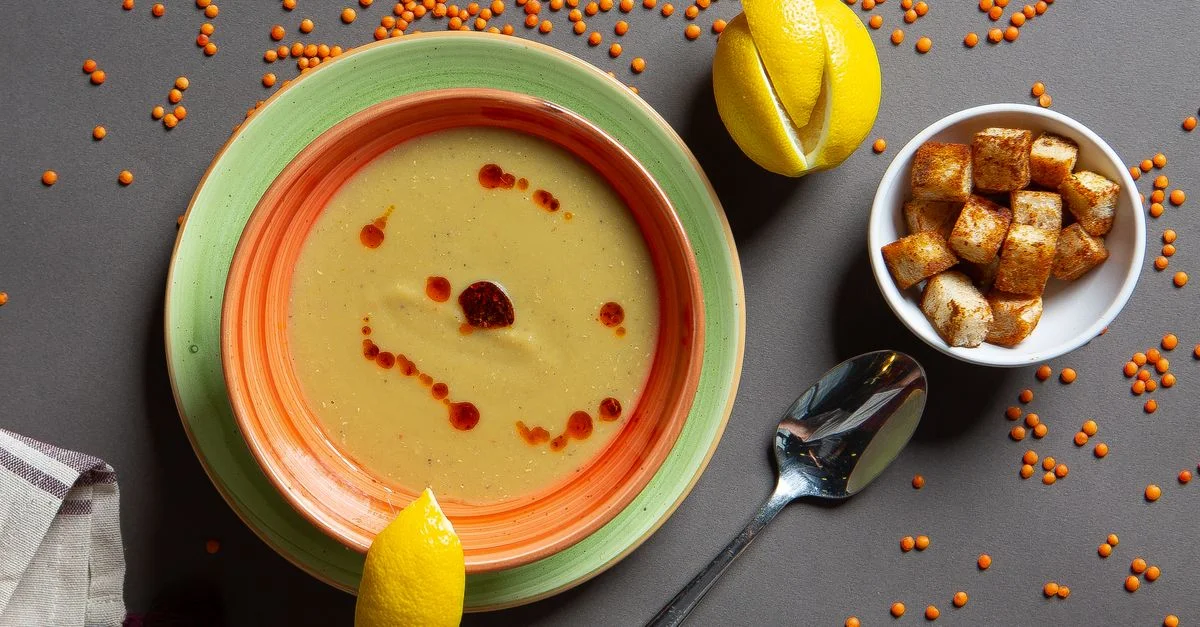 Un tazón caliente de sopa de lentejas rojas servido con rodajas de limón y picatostes, presentado como una composición cenital.