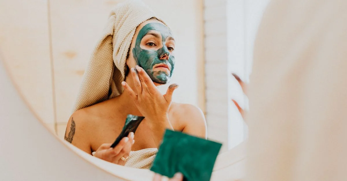 Una mujer se aplica una mascarilla facial de arcilla verde en el espejo del baño, promoviendo el autocuidado y las rutinas de cuidado de la piel.