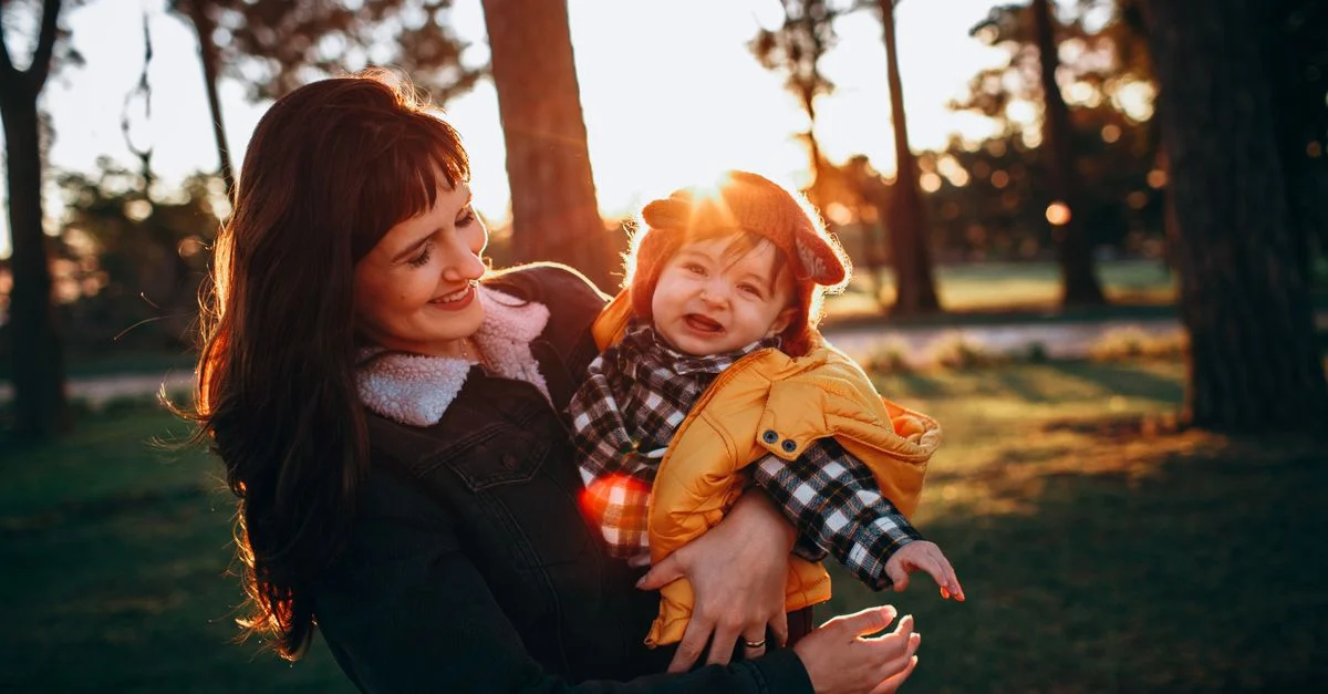 Una madre feliz sosteniendo a su bebé en un parque soleado durante el atardecer, capturando un momento cálido y alegre.