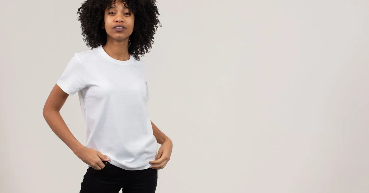 Joven mujer con cabello rizado posando con confianza en una camiseta blanca sobre un fondo liso.
