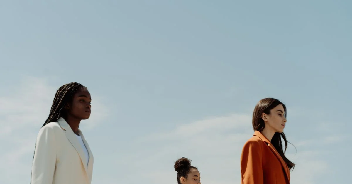 Tres mujeres a la moda en blazers elegantes, de pie con confianza bajo un cielo despejado.