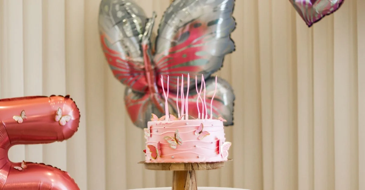 Tarta de cumpleaños rosa con decoraciones de mariposas, rodeada de globos en interiores.