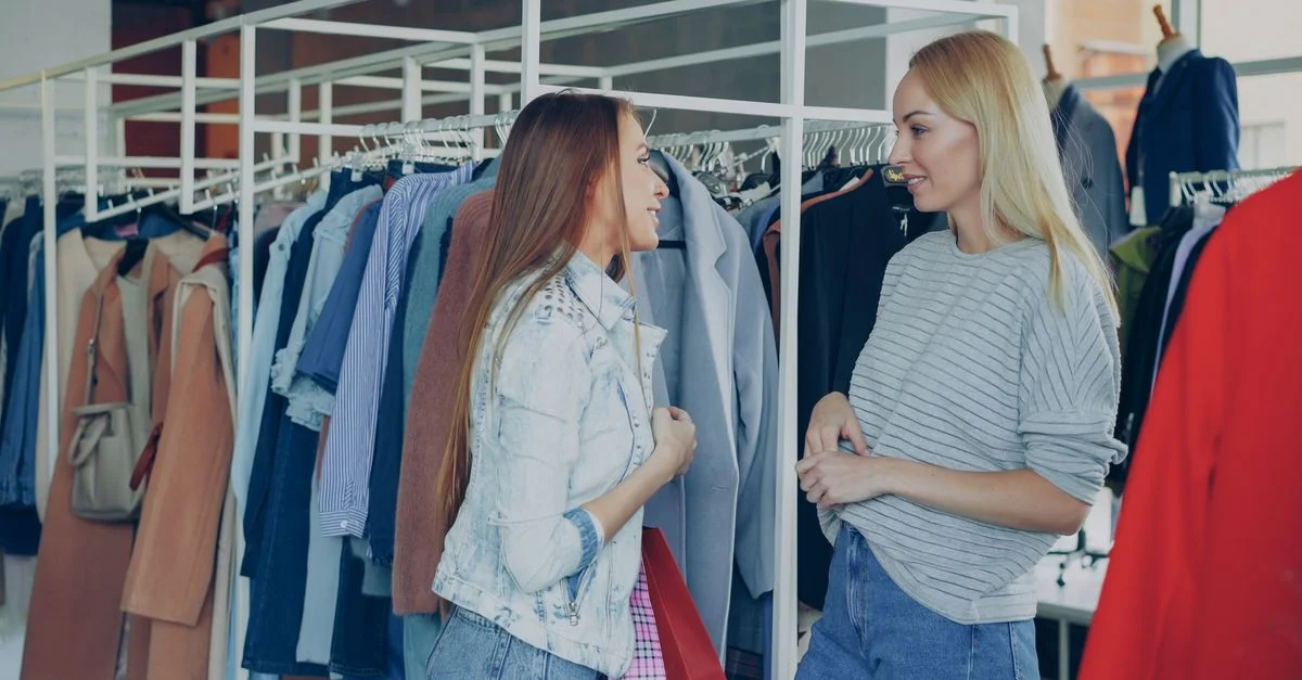 Dos mujeres charlando entre percheros de ropa en una elegante tienda de moda, compartiendo consejos de compra.