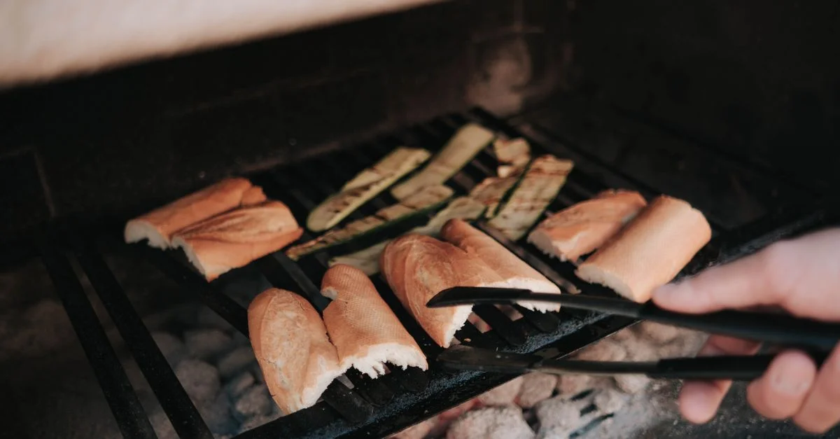 Vista en primer plano de pan y verduras asándose en una barbacoa con pinzas.