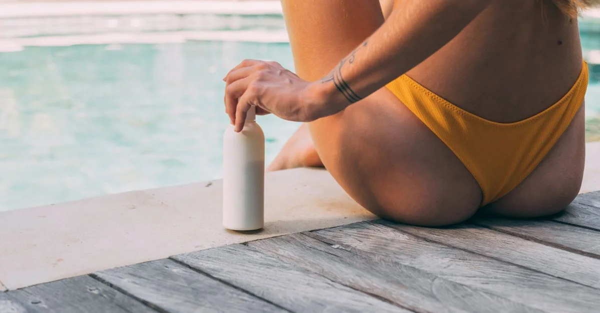 Una mujer sentada al borde de la piscina, sosteniendo una botella de protector solar bajo la luz del sol de verano.