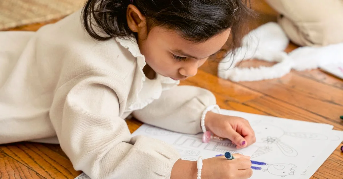 Una niña joven está coloreando creativamente en un libro mientras está tumbada en un suelo de madera en casa, concentrándose intensamente.