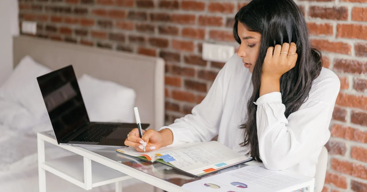Una joven estudia en interiores con un portátil, escribiendo notas en un cuaderno sobre un escritorio.