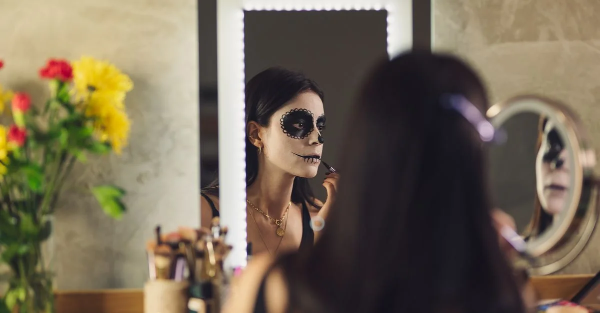 Mujer aplicando maquillaje de Catrina para el Día de los Muertos frente a un espejo adornado con flores.
