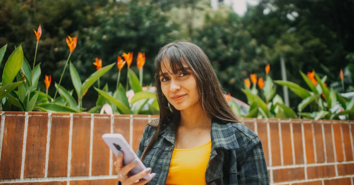 Mujer con una blusa amarilla utilizando un smartphone, rodeada de flores vibrantes en Guatemala.