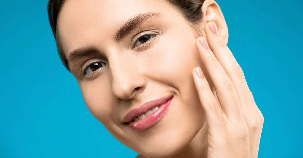 Primer plano de una mujer sonriendo con maquillaje natural sobre un fondo azul, mostrando belleza y cuidados de la piel.