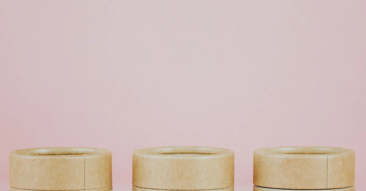 Tres tarros de cosméticos ecológicos con etiquetas botánicas sobre un fondo rosa.