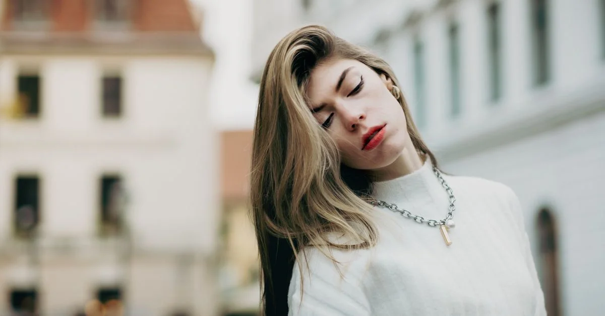 Mujer elegante en suéter blanco posando con los ojos cerrados en un fondo urbano.