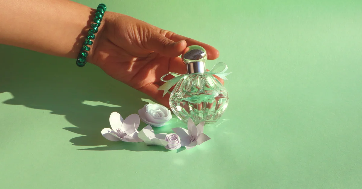 Una mano con pulsera sostiene una botella de perfume rodeada de flores de papel sobre un fondo verde.