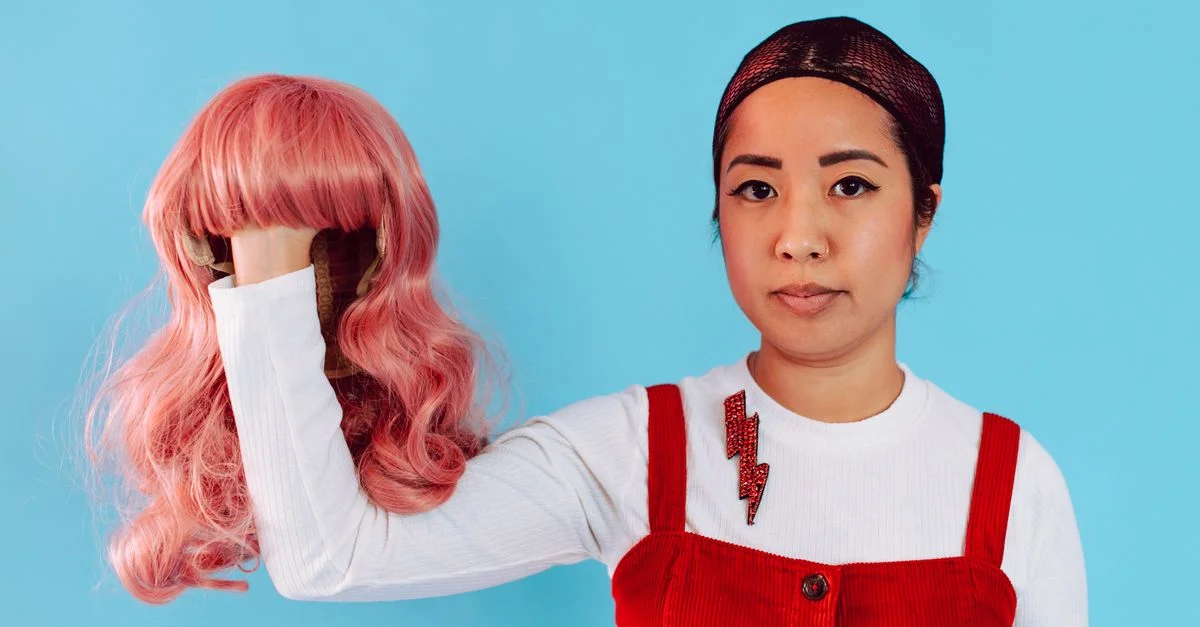 Mujer asiática segura una peluca rosa, vestida con overoles rojos, de pie frente a un vibrante fondo azul.