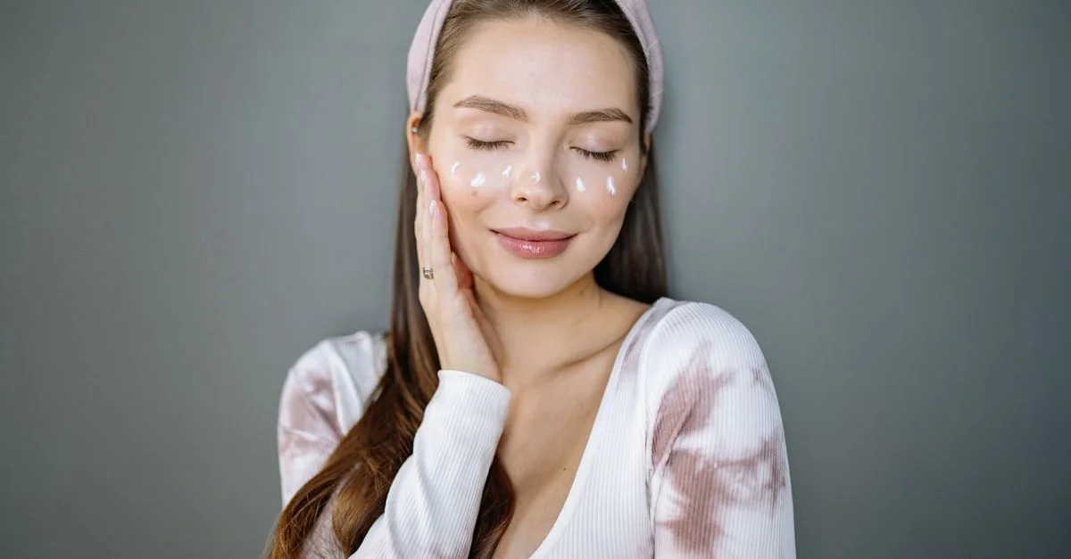 Retrato de una mujer sonriente con crema facial, ojos cerrados y mano en la mejilla sobre un fondo gris.