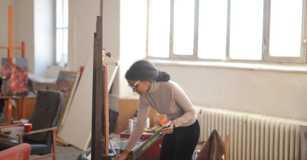 Una mujer pintando en un estudio de arte bien iluminado, rodeada de lienzos y herramientas.
