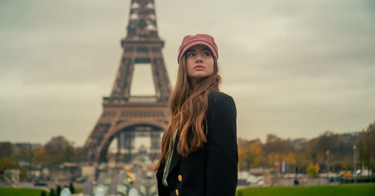 Una joven mujer se encuentra elegantemente en un parque de París con la Torre Eiffel de fondo, disfrutando de la ambientación otoñal.