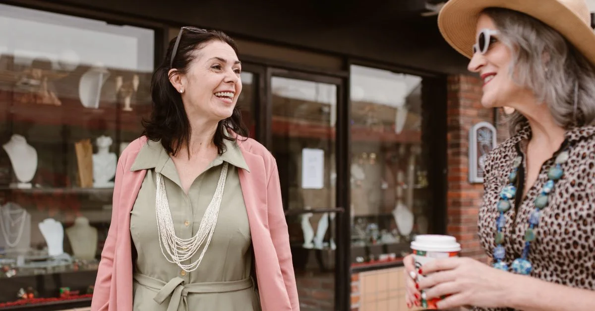 Dos mujeres caucásicas sonriendo y charlando al aire libre, una sosteniendo una taza de café.