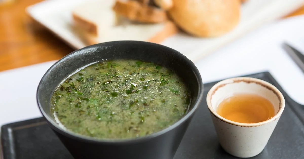Un cuenco de sopa verde herbal servido con pan y un dip al lado, ofreciendo una comida deliciosa.