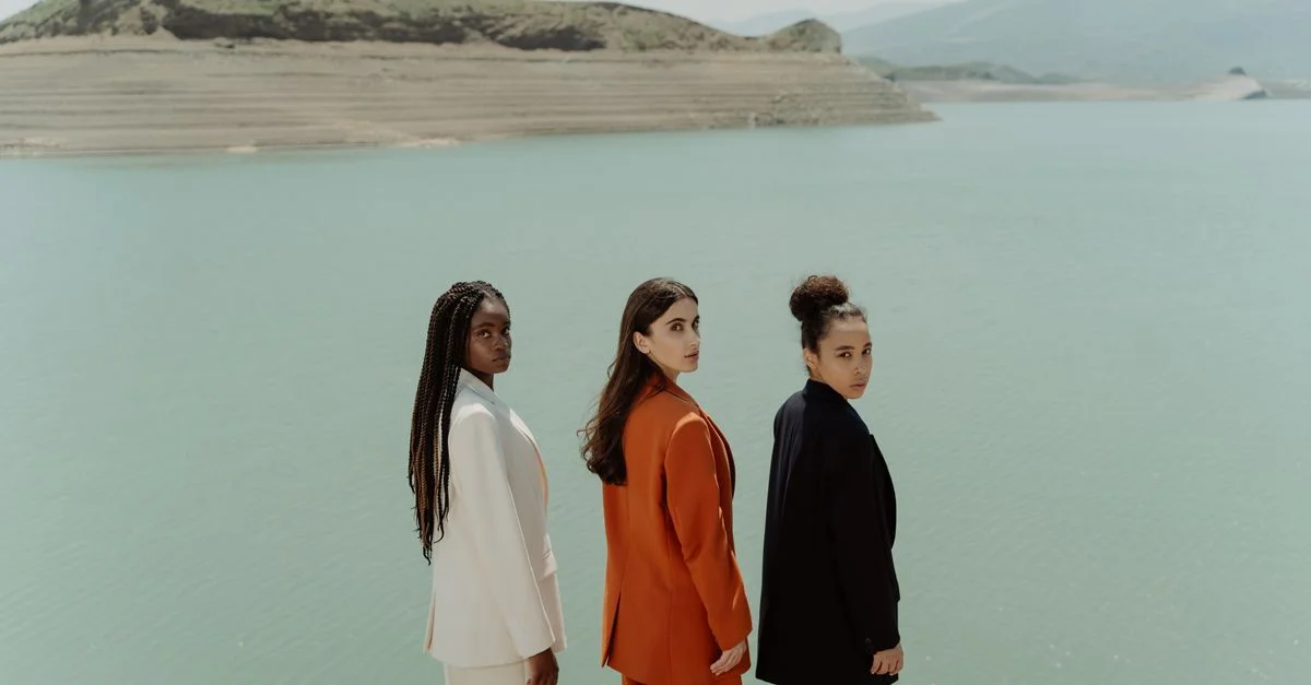 Tres mujeres en blazers coloridos están elegantemente junto a un lago tranquilo, desprendiendo moda y diversidad.