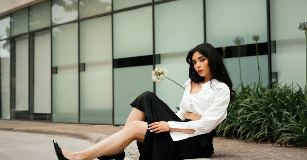 Mujer elegante vestida de blanco y negro sentada frente a la fachada de un edificio moderno, luciendo moda urbana.