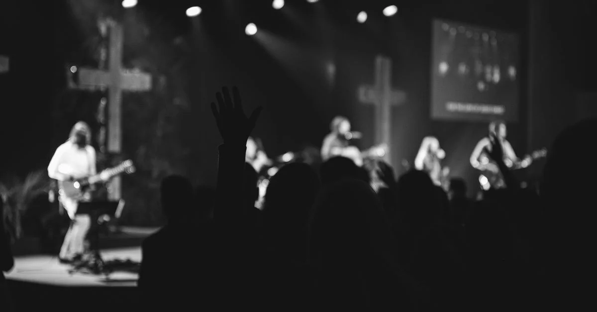 Audiencia silueteada en un concierto con músicos en el escenario y cruces al fondo.