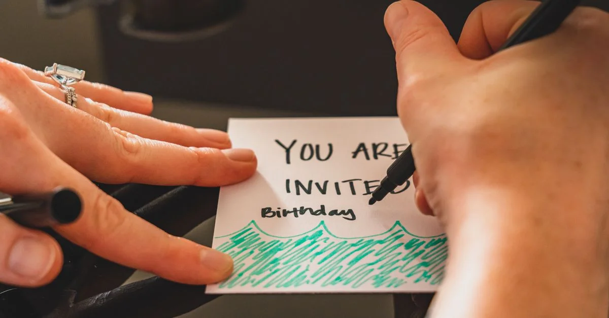 Manos escribiendo una tarjeta de invitación de cumpleaños personalizada utilizando un marcador sobre papel.