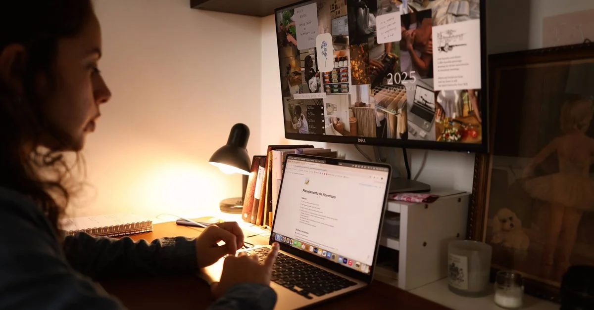 Una mujer trabaja en el escritorio de su oficina en casa con una computadora portátil y un monitor bajo una luz cálida.