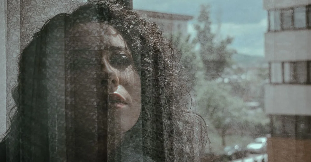 Una mujer con cabello rizado mira pensativa a través de una ventana reflectante, capturando un ambiente sereno y reflexivo.