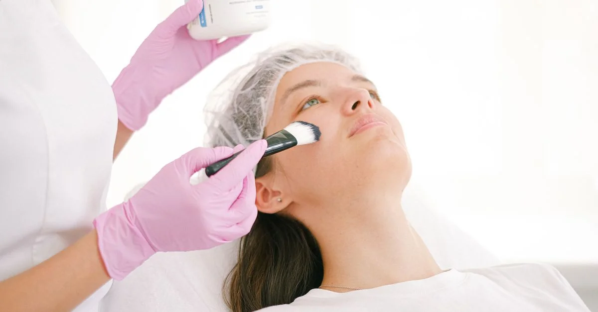 Una mujer recibiendo un tratamiento facial en una clínica por un profesional utilizando un cepillo y ungüento.